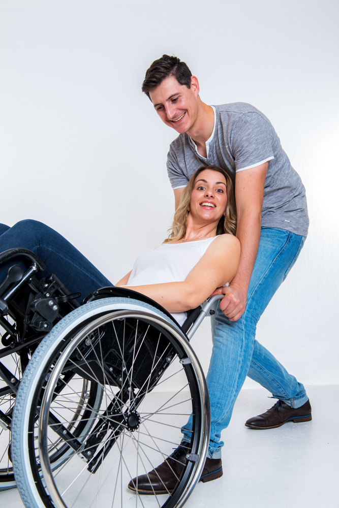 A white man with short brown hair in blue jeans and a gray t-shirt smiles at a blonde woman in a manual wheelchair as he tips her chair backwards from behind.