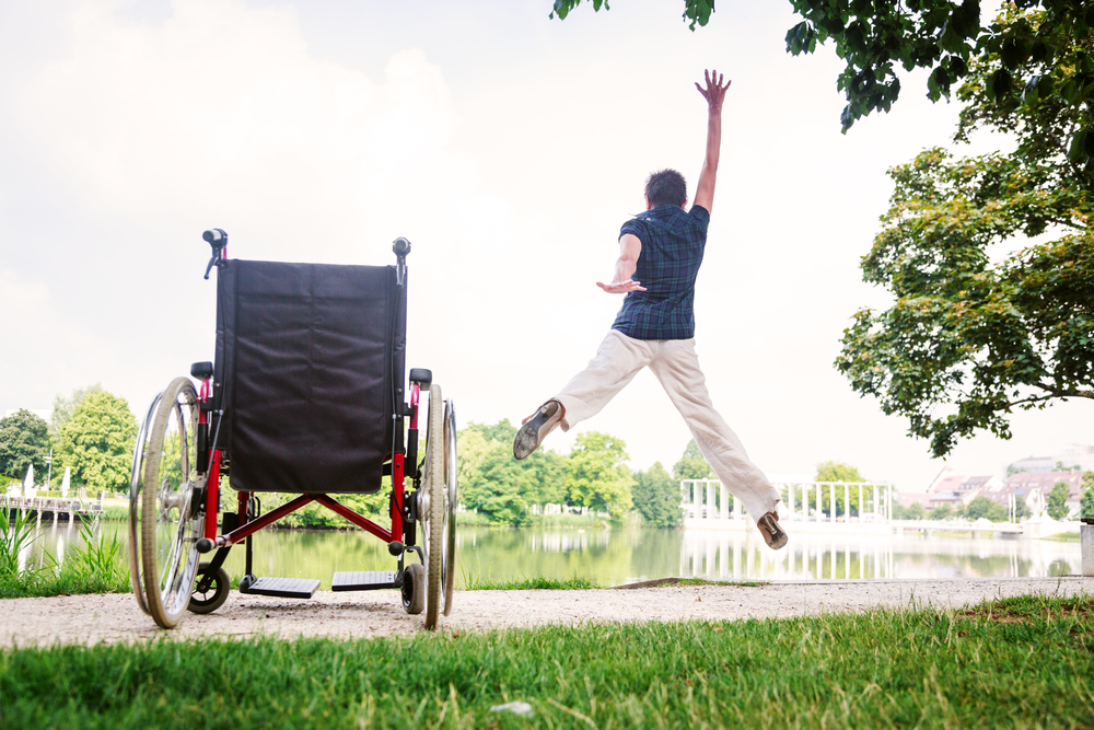 A woman with short brown hair in a blue short sleeved shirt and khaki pants leaps out of a manual wheelchair with her arms up in the air. There is a large pond in the background.