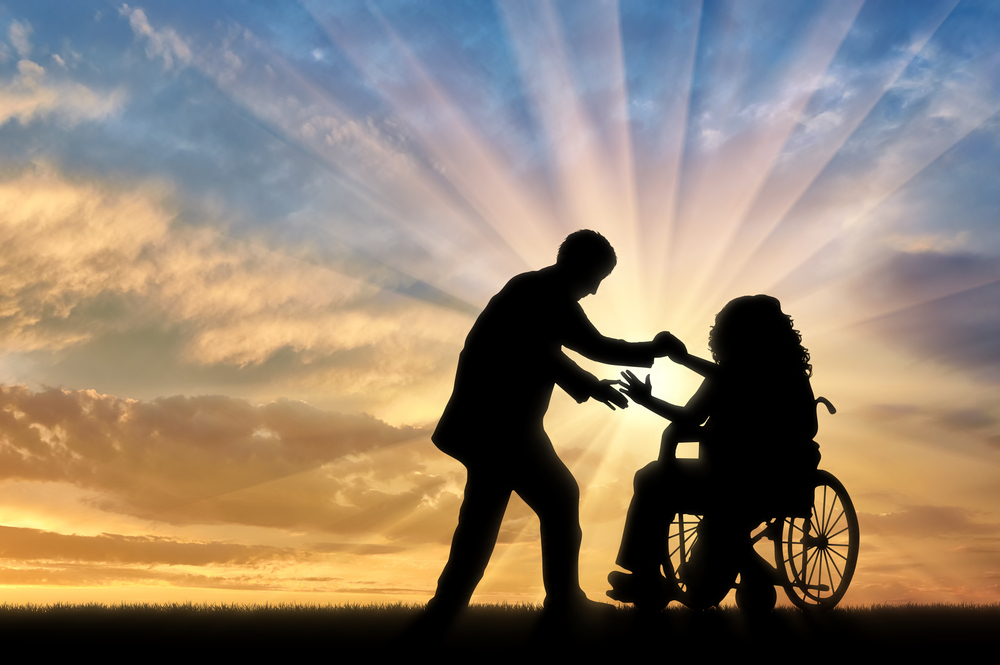 In silhouette against a sunset background, a man reaches out to take the hands of a woman in a manual wheelchair.