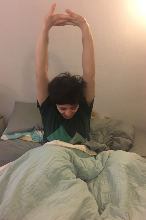 A white woman with short brown hair wearing a dark green t-shirt sits up in bed stretching her arms above her head. She's under a light green blanket, and the bedsheets are gray.