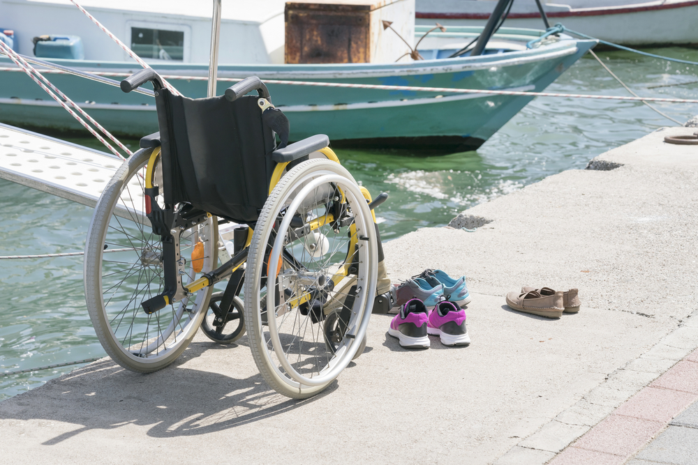 A manual wheelchair with a yellow frame, black seat, and gray tires sits unoccupied on a dock next to the water. Three pairs of shoes sit next to the chair. There are no people in the photo, only a boat in the background.