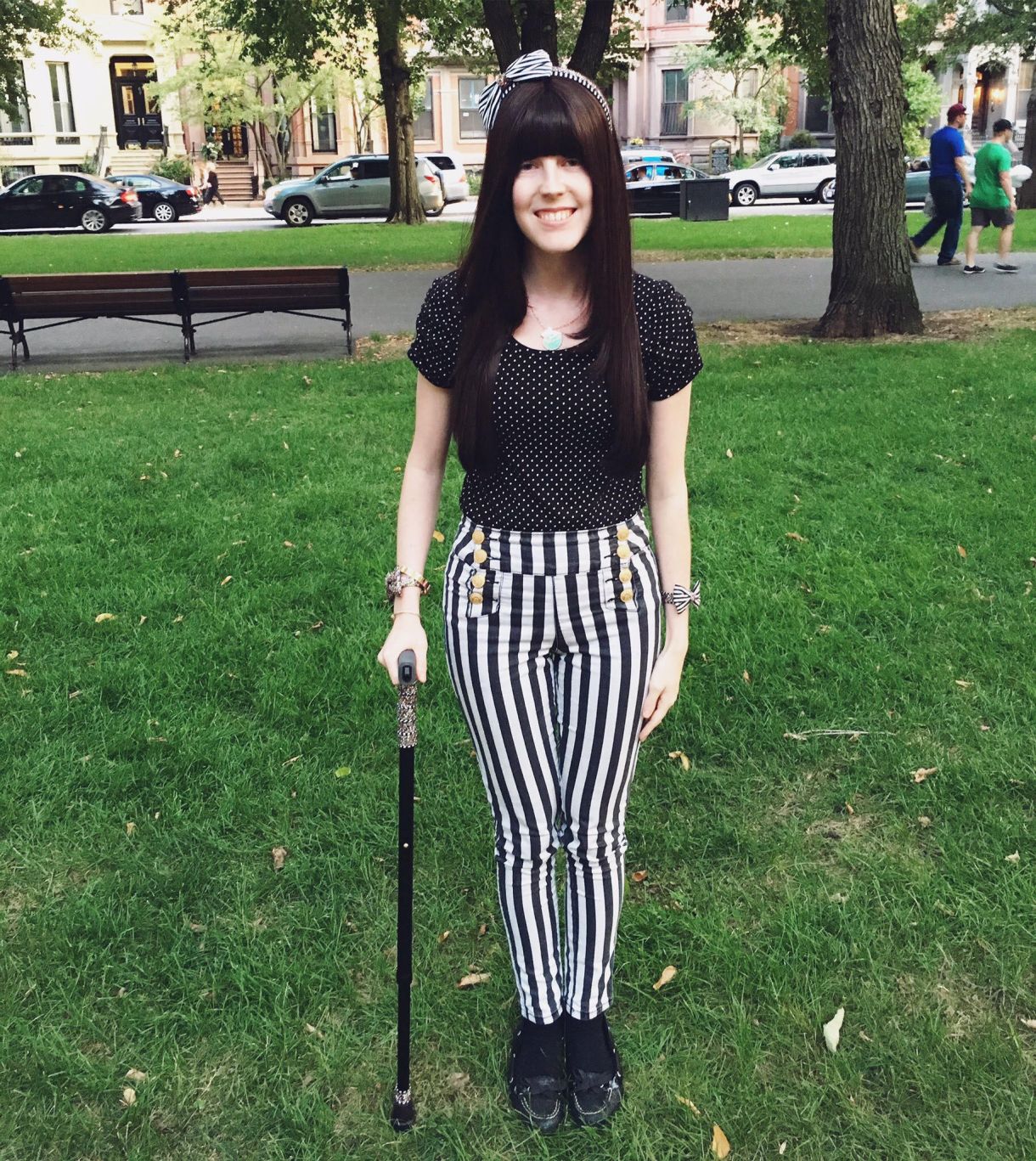 A white person in her twenties with long brown hair and blunt bangs stands on the grass in a park. She's holding a black cane and wearing black and white striped pants and a black top with small white polka dots, plus a headband with black and white stripes and a bow.