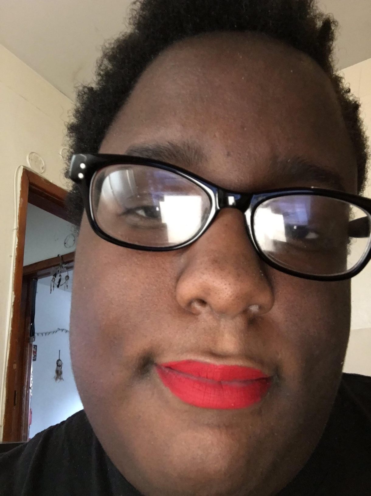 Selfie of a black woman with short black hair and wearing red lipstick and black square-framed glasses smiling close-mouthed into the camera.