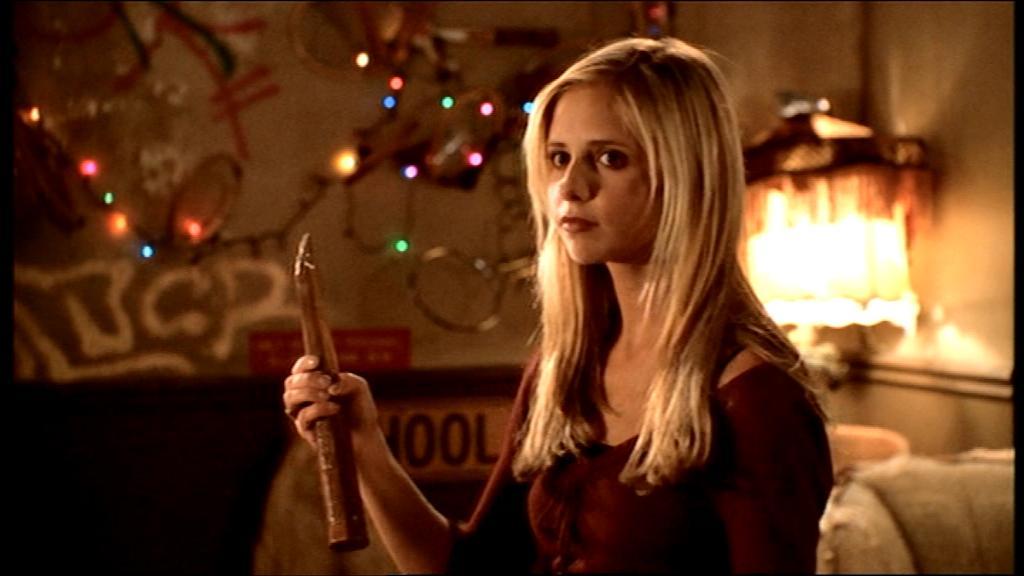 Buffy Summers, a slayer of vampires, holds a stake. She's inside a house that has weird Christmas lights and lamps and signs and stuff.