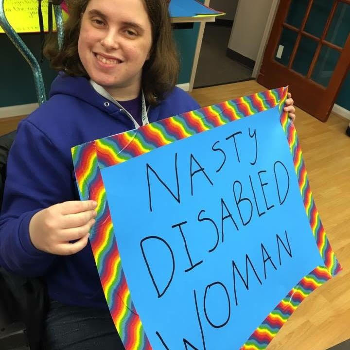 A white woman in her mid twenties with brown hair and wearing a blue hoodie holds up a blue sign with a rainbow border that says "Nasty Disabled Woman."