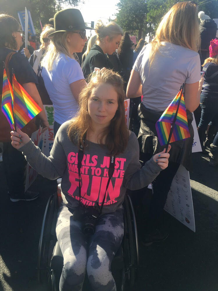 A white teenage woman with shoulder-length blonde hair sits in her wheelchair holding small rainbow flags in each hand. Her shirt says "Girls just wanna have fundamental rights" in hot pink letters.