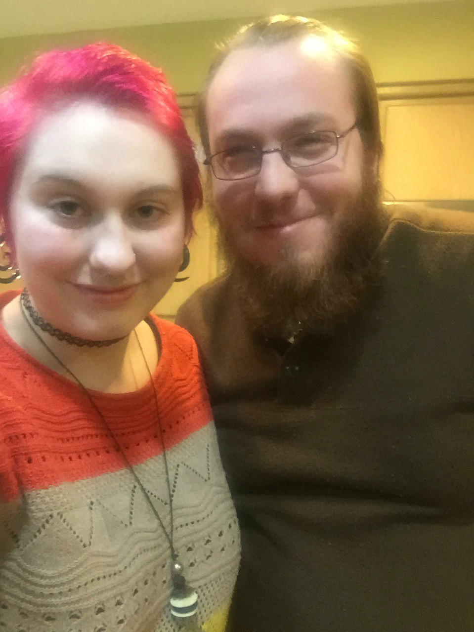 Kaety, a pale-skinned person with pink hair and wearing an orange and gray sweater, stands next to their fiance Matt, a pale-skinned man in a dark sweater with a brown beard.