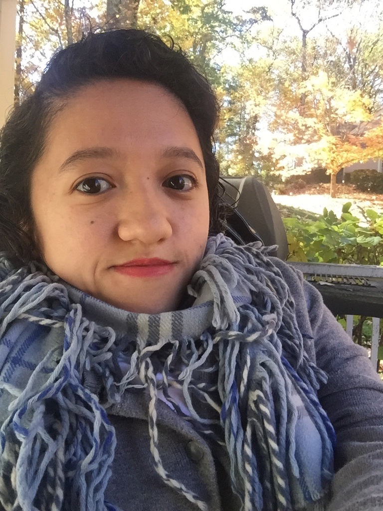 An Asian woman in her late twenties sits outside in her electric wheelchair, looking at the camera. She has black hair and red lips and is wearing a gray plaid scarf with tasseled ends.