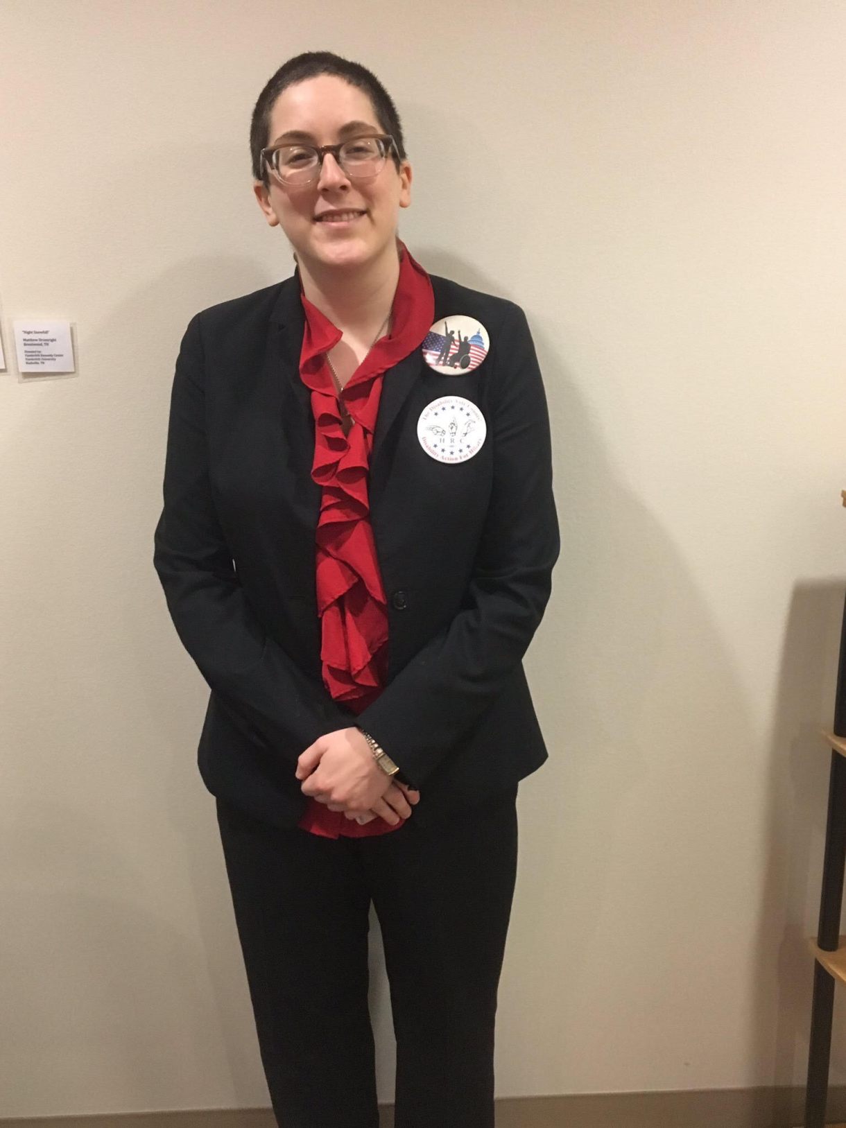 A white woman in her twenties with short brown hair stands with her arms folded at her waist against a white wall. She's wearing a black pantsuit with a red ruffled shirt underneath.