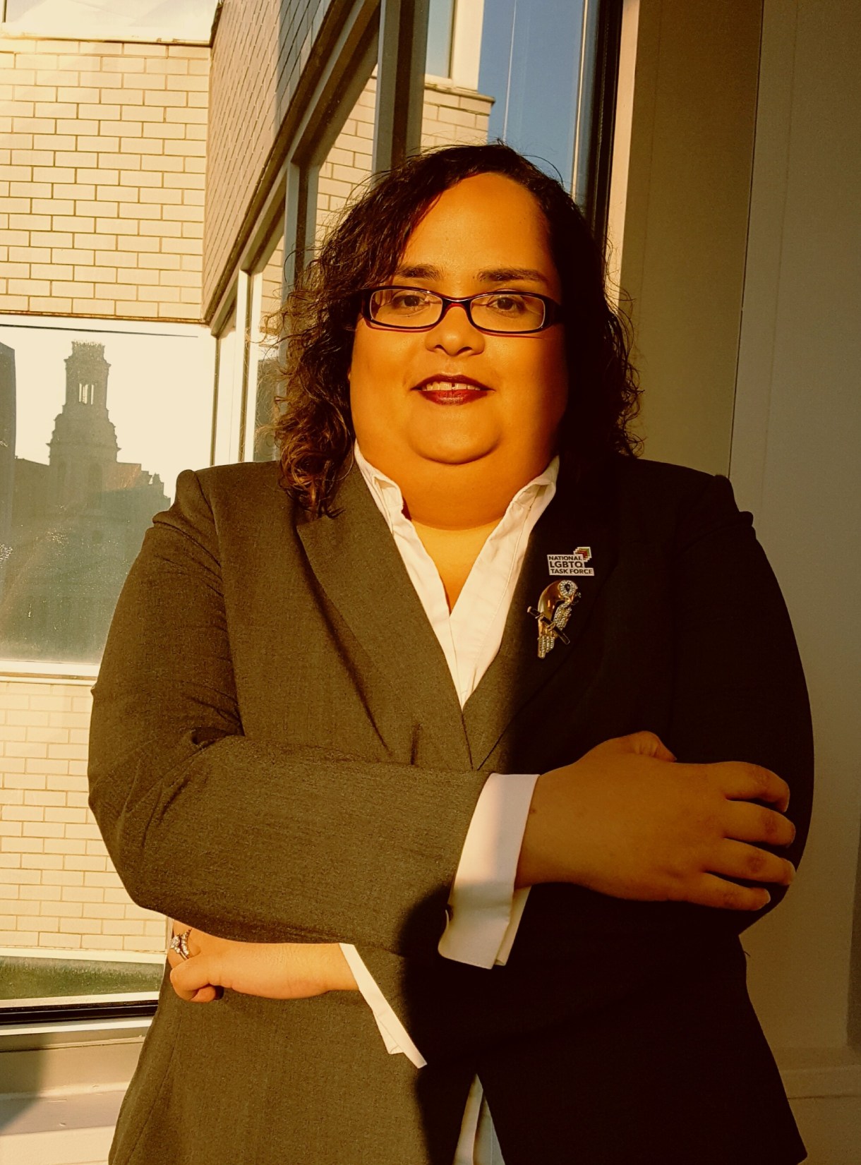 A Latina woman with square eyeglasses and red lips wearing a white shirt and black blazer stands with her arms crossed in front of a white brick building. The sun is lighting up her face and the right half of her body; her left arm is in shadow.
