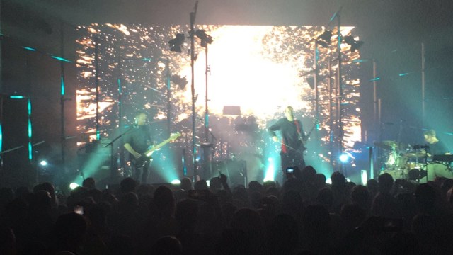 A three-piece band (bassist on the left, lead singer and guitarist in the middle, drummer on the right) play onstage in front of an explosion of bright white light. The crowd is reaching up to them from the floor, and the guitarist is playing his instrument with a violin bow instead of a pick.