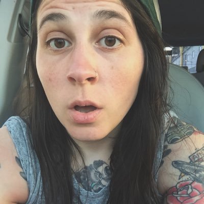 A white woman with long brown hair, brown eyes, and a nose ring, with her chest and upper arms covered in tattoos.