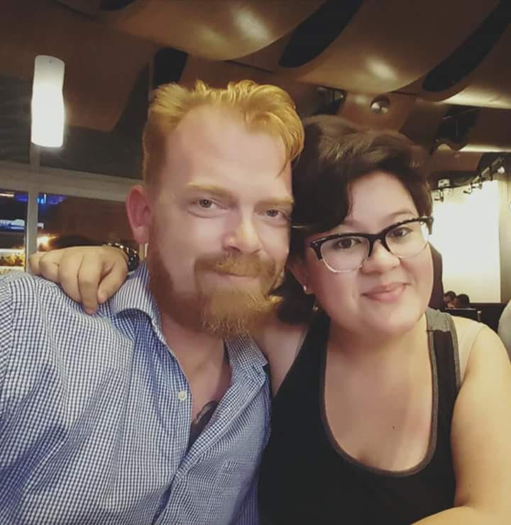 A young couple (a white man with red hair and a Latinx person with dark brown hair and glasses) smile at the camera with their arms around each other's shoulders.