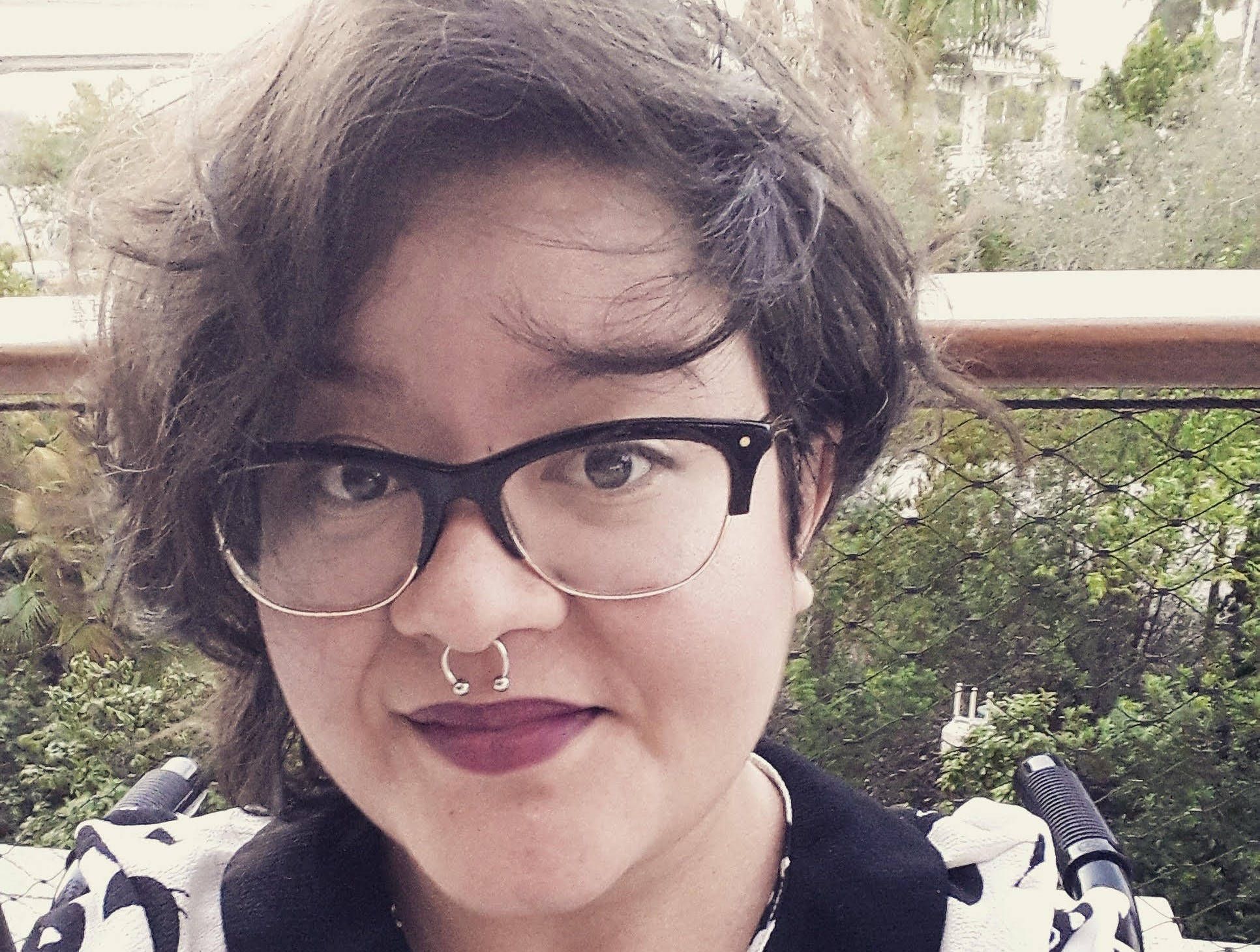 A Latinx person with short black hair wearing red lipstick, black framed glasses, and a pin that says "PRIDE" in rainbow letters smiles into the camera.