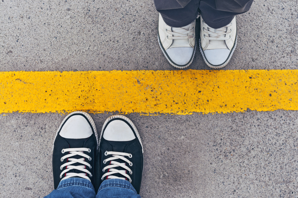 An overhead view of two pairs of feet in sneakers, each standing on either side of a thick yellow line.