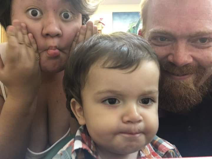 A family of three, two parents and their young child, smile into the camera. The mom, who's on the left side of the frame, is making a fish face.