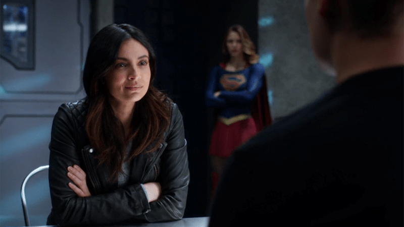 Maggie sits at the table, Supergirl stands watch behind her 