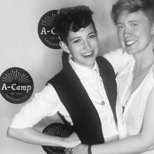 Two women in formal wear pose with their arms around each other and smile for the camera in front of an A-Camp branded backdrop.