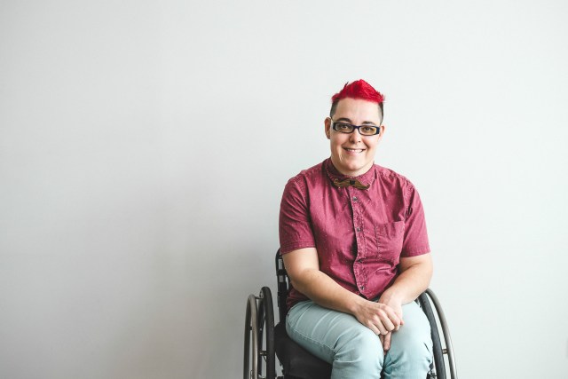 A woman with a red mohawk wearing jeans and a reed shirt sits in her wheelchair against a blank wall, smiling.