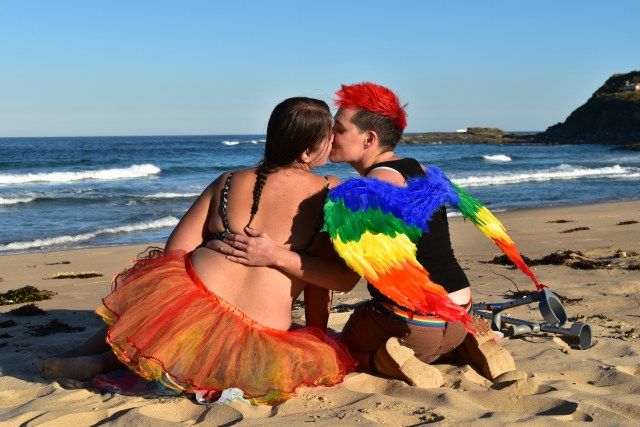Two women sit next to each other on a beach, kissing. One is wearing a set of rainbow angel wings, the other an orange skirt.