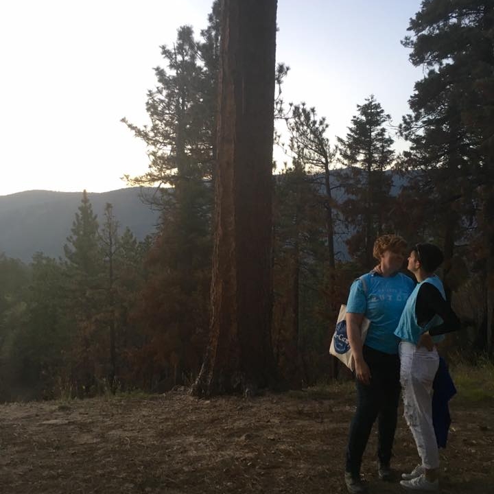 Two women look lovingly at each other while standing on a wooded mountaintop. They are both wearing light blue shirts, and one is carrying an A-Camp tote bag.