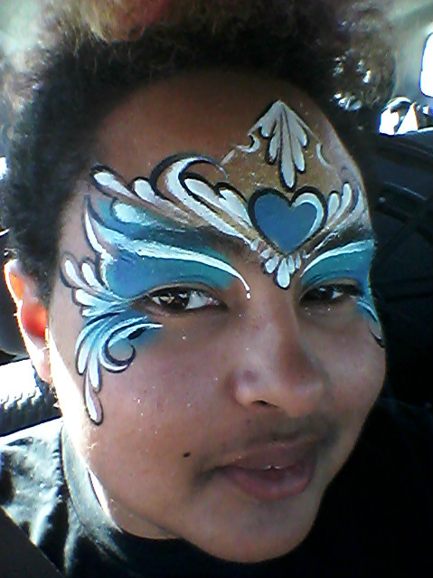 A young person with brown skin and dark hair looks into the camera. Their face is painted blue and white over the eyes, with a heart in the center of their forehead.