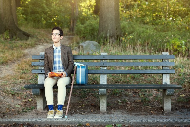 A dapper-looking person with a wooden cane sits at the end of a park bench.