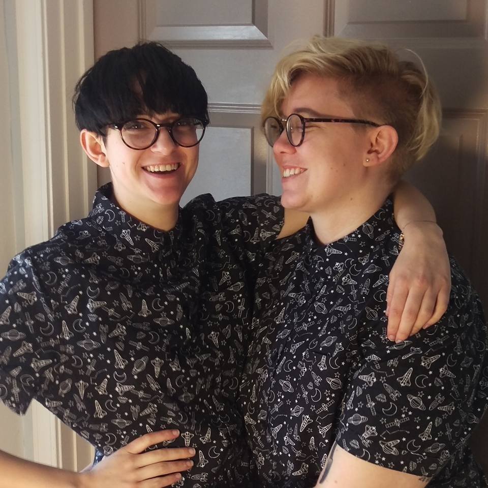 A couple standing with their arms around each other, both smiling — one at her partner, one at the camera. They are wearing matching short-sleeved button down shirts, patterned with outlines of planets, rocket ships, and stars.