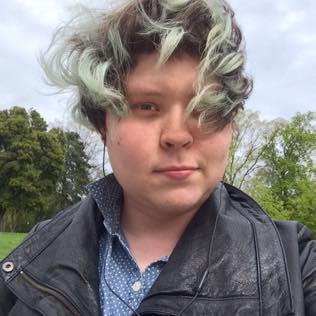 A nonbinary trans girl with short brown hair that is faded green at the tips looks into the camera. She is standing outside and the wind has blown her hair over into her eyes.