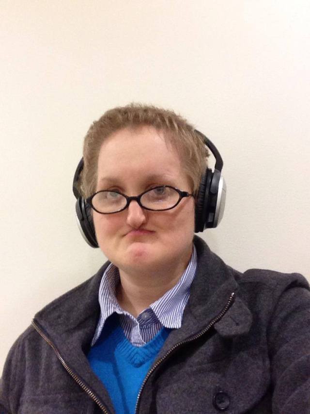 A young white person with short blond hair wearing dark roundish glasses, wearing a dark blue pinstriped white collared shirt, a bold blue sweater, and a dark gray woolen coat, standing against a white wall.