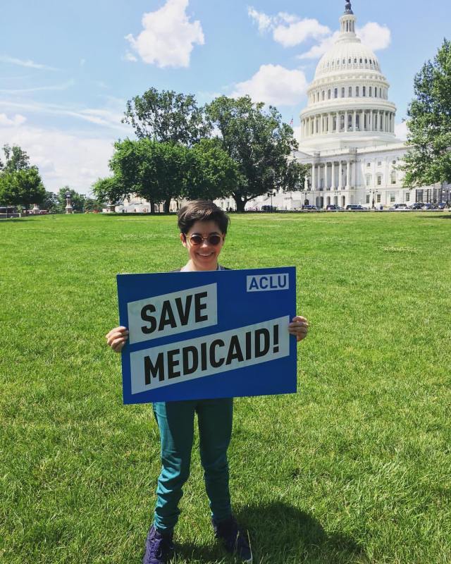 A woman with short brown hair wearing sunglasses and teal jeans stands in front of the U.S. Capitol Building holding a blue and white sign that says "Save Medicaid!".