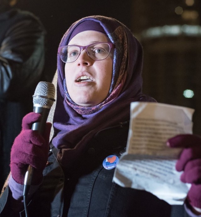 Mahdia Lynn in the process of giving a speech. She is wearing purple glasses, purple gloves, and a purple head scarf with a black coat, holding a microphone in one hand and a piece of paper in the other.