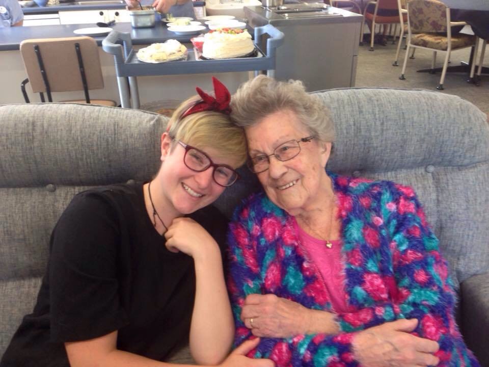 Scout sits on a couch next to their grandmother, and they both smile for the camera. Nana is wearing an amazing floral printed jacket, and Scout has a vibrant red bow headband on.