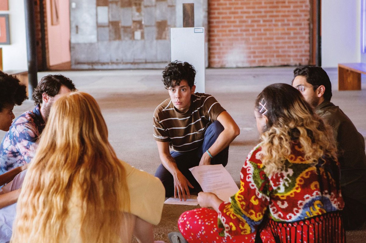 Image: An art gallery in Marfa. Devon, played by Roberta Colindrez, is presenting her play to a group of artists who are sitting in a semi-circle around her. She is wearing a brown t-shirt with white stripes and has dark, curly hair. One of her hands is on a piece of paper on the floor, the play script. Wee see the backs of five students circled around her. 
