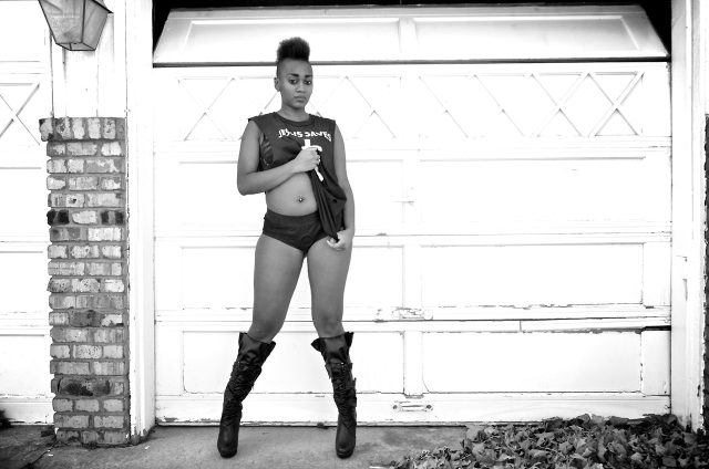 @nikia_deshawn in knee-high boots, tanktop, and harness, posing against a garage door