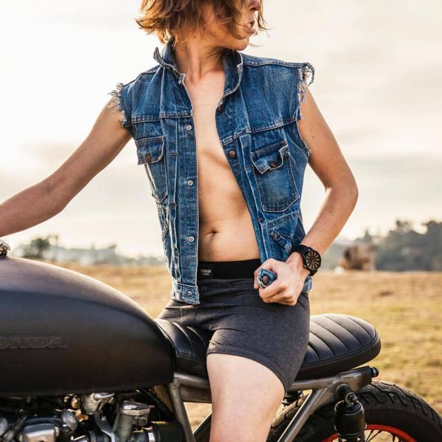 @pancha88 straddling a motorcycle in denim vest, no shirt, and grey strap-on harness