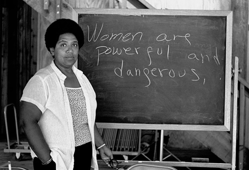 Audre Lorde stands next to a chalk board with the phrase "Women are powerful and dangerous" written on it