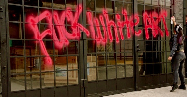 Marisol spray paints "Fuck White Art" on gallery wall.