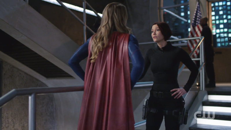 Supergirl has her hands on her hips, Alex has one hand on her hip one on the rail