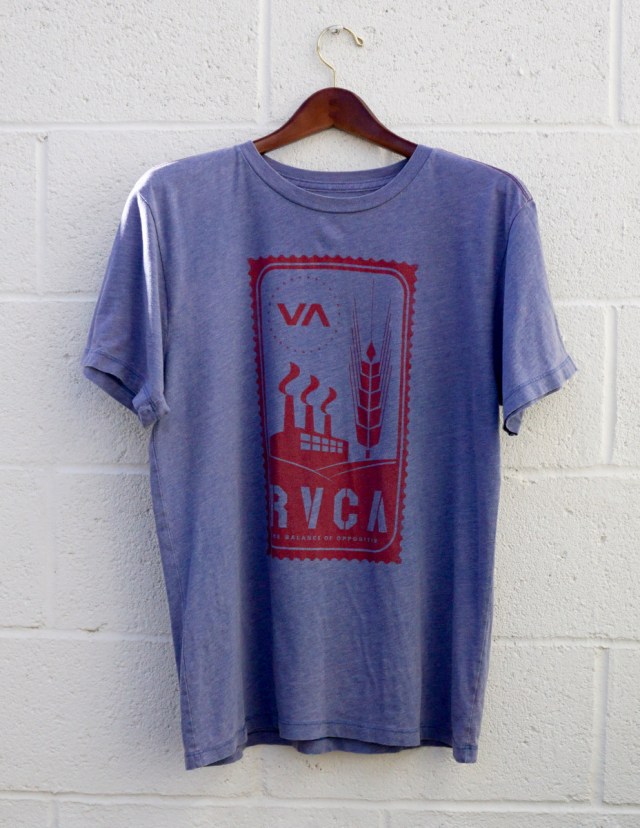 A blue t-shirt with a red graphic design, with short sleeves, hanging against a white brick wall.