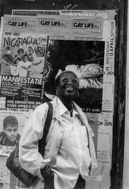 A black and white photo of Angela Bowen standing in front of a wall that's covered in posters and flyers, apparently for local art and queer community. She's wearing a white button down shirt, white jacket, large glasses, and a bag over her shoulder. She's smiling up toward the sky at something off-camera.