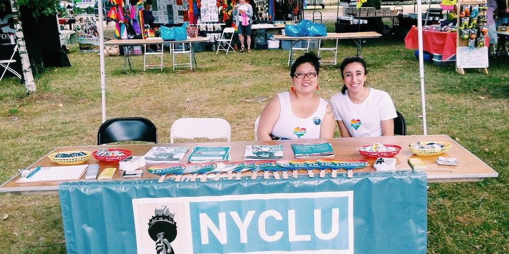 Two people in rainbow heart t-shirts sit at a table with a banner across the front that reads, "N-Y-C-L-U: New York Civil Liberties Union" and literature, fans, and pins at a Pride event with more tables and people in the background.