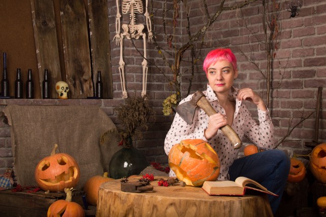 A pink-haired femme-ish woman holding an axe, surrounded by pumpkins, and looking off into the middle distance, with a book on the treestump in front of her for some reason (she is also inside)