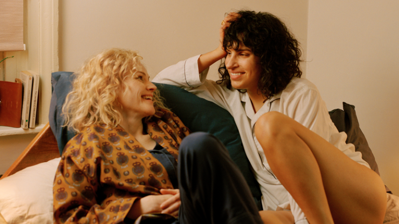 Image: Leila (played by Desiree Akhavan), an Iranian-American woman with short dark hair in a pajama shirt, is on a bed looking lovingly at her girlfriend, an older white woman with curly blonde hair and a robe.