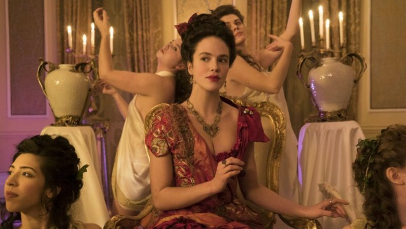 Image: a fancy assortment of women at a brothel in long-ago England. Charlotte is front and center in a fancy red dress, and women are posing behind her like they are art. Harlots is one of the lesbian shows on Hulu.