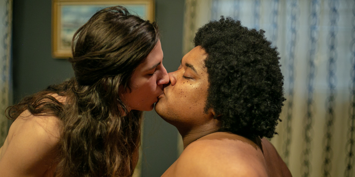 A black bearfemme with shortish natural hair and bare shoulders and a white trans woman with long brown hair and bare shoulders make out in this Crash Pad Series still