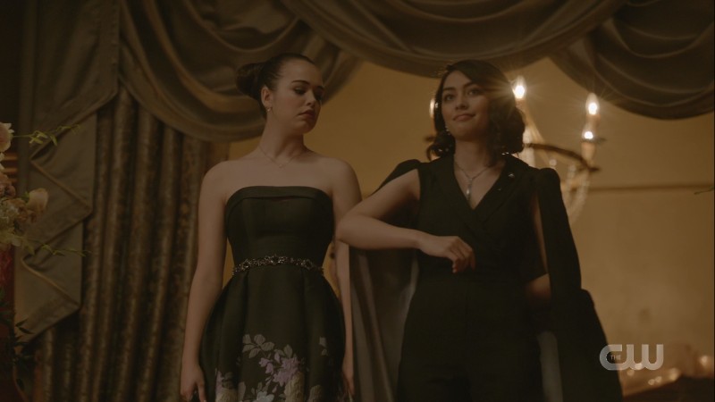 Penelope in a black jumpsuit with a cape escorts Josie in a black dress down the stairs