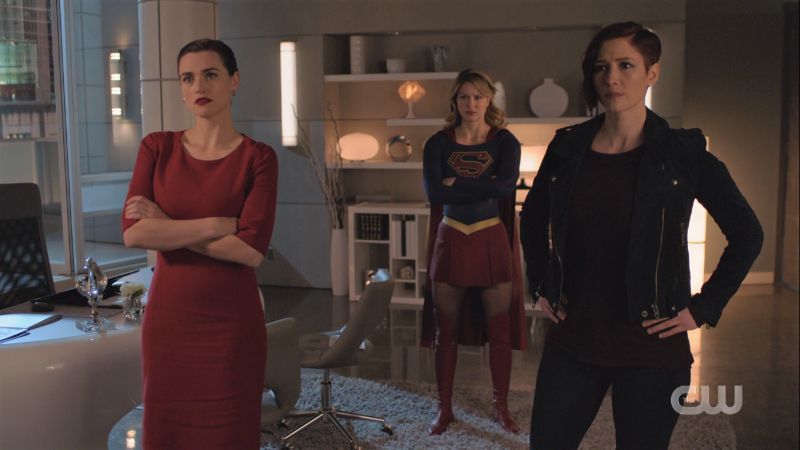 Lena Supergirl and Alex sand around with crossed arms or hands on hips staring angrily at the TV