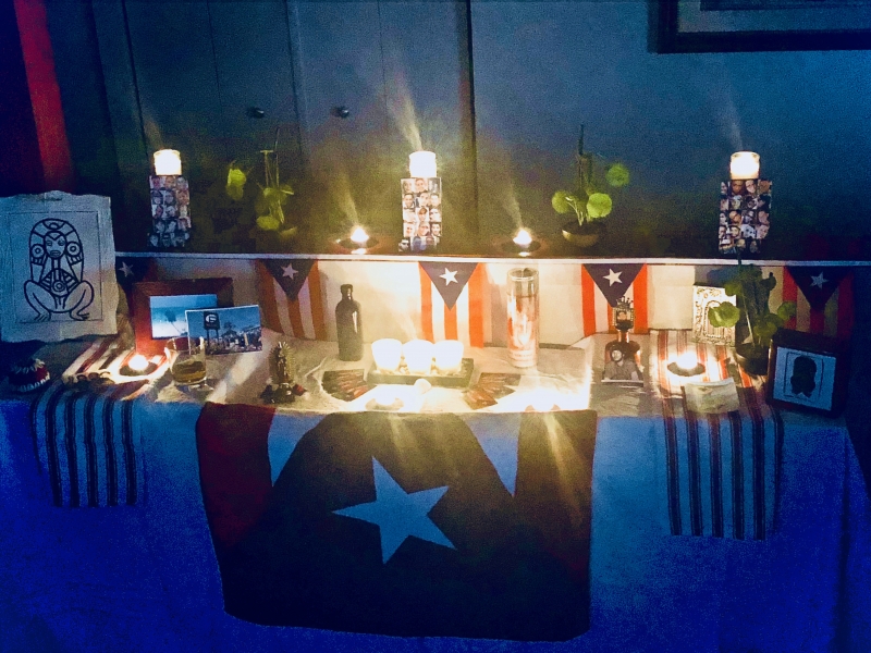 A home photograph of an altar built in a living room with a collection of white table cloth, candles, and Puerto Rican flags. The photograph is in the dark with candles lit.
