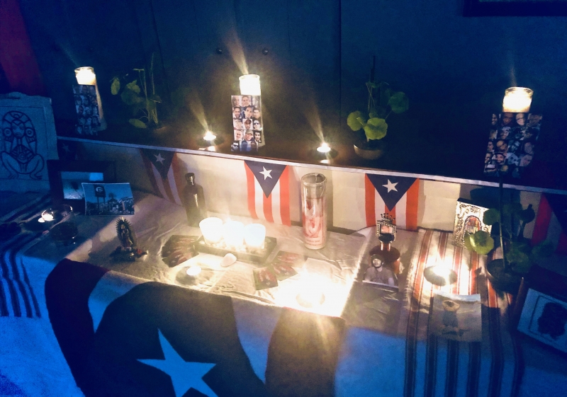 A home photograph of an altar built in a living room with a collection of white table cloth, candles, and Puerto Rican flags. The photograph is in the dark with candles lit.